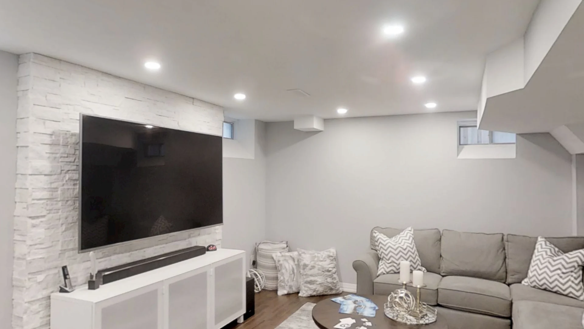 A finished Harmony basement seen from the middle of the room: the white stacked-stone TV wall, the grey sectional and the pot-lit ceiling
