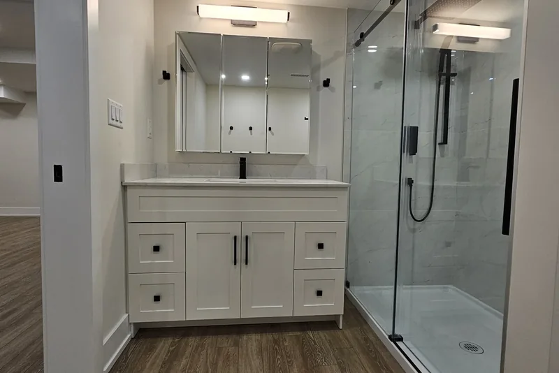 Finished three-piece basement bathroom in Vaughan seen from the doorway: a white shaker vanity with a quartz top and a mirrored cabinet above it, and beyond it a marble-look tiled shower closed with a black-framed sliding glass door, on vinyl plank flooring.