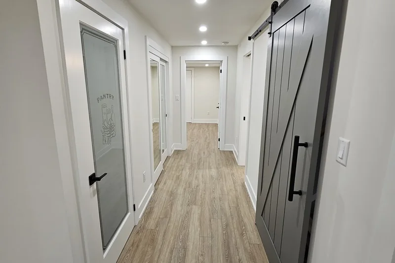 Finished basement hallway cost, Vaughan project: a grey Z-brace barn door on a black track fills the right-hand side, an etched glass door marked PANTRY stands on the left, and vinyl plank flooring runs under a line of pot lights to the doors at the far end.