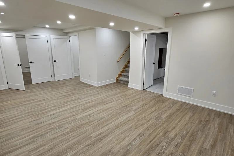 Finished basement in Vaughan looking across the open floor: a bank of white doors on the left, a staircase rising behind its wall in the centre, and an open white door on the right showing the unfinished service room.
