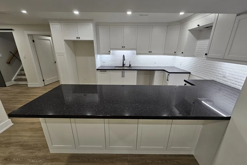 Basement finishing cost — Finished basement kitchen in Vaughan seen across the peninsula: white shaker upper and base cabinets, a white subway tile backsplash lit by under-cabinet lighting, black speckled stone counters running in a U with a black tap over the sink, and the foot of the staircase visible through an opening at the far left.