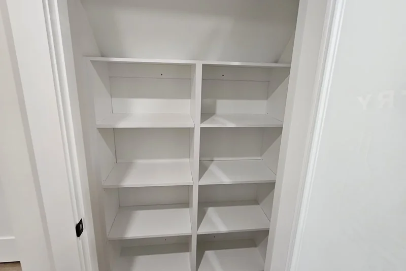 Basement finishing cost — The finished pantry under the staircase in a Vaughan basement: white melamine shelves in two bays with the underside of the stair sloping across above them, vinyl plank flooring at the bottom, and the etched glass door standing open at the right.
