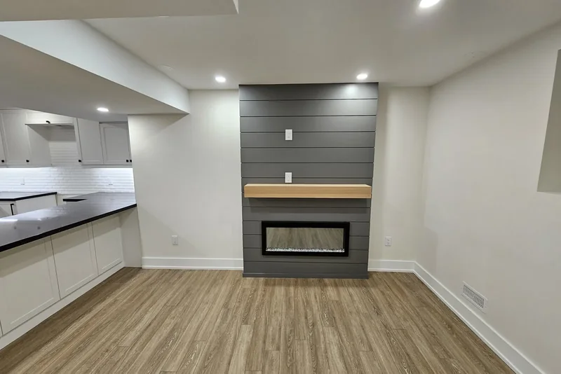 Finished basement family room cost, Vaughan project: a dark grey shiplap chimney breast carrying a light oak floating mantel above a linear electric fireplace, wide plank flooring across the room, and the white bar cabinets and black counter at the left edge.