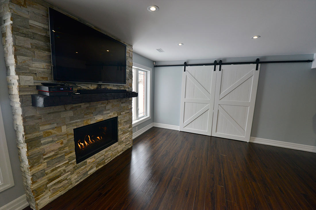 Basement fireplace in Aurora: a natural stone wall, a rustic mantel and white barn doors, by Harmony Basements